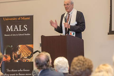 A man in a University of Miami stole speaks to a class of MALS alumni. 